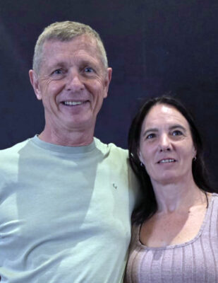 A man in a light green shirt and a woman in a light pink top stand together, smiling, against a dark background, conveying a warm and friendly atmosphere.