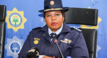 A South African police officer in uniform sits at a podium with microphones, delivering a speech. The South African Police Service logo is in the background.