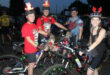 Four cyclists at night wearing festive attire and light-up helmets, with decorated bikes. A joyful atmosphere with Christmas lights and holiday cheer.