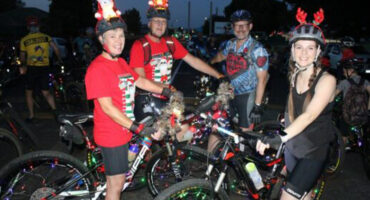 Four cyclists at night wearing festive attire and light-up helmets, with decorated bikes. A joyful atmosphere with Christmas lights and holiday cheer.