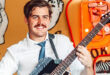 Smiling Marco van Deventer with a mustache plays a guitar indoors, wearing a white shirt and tie. Bright, artistic background with bold, cartoon-like designs.