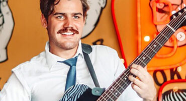 Smiling Marco van Deventer with a mustache plays a guitar indoors, wearing a white shirt and tie. Bright, artistic background with bold, cartoon-like designs.