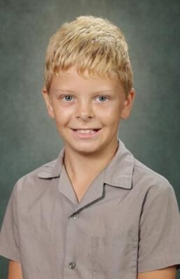 A young boy with blonde hair smiles warmly against a gray studio background. He wears a light gray short-sleeved shirt, conveying a cheerful, friendly tone.