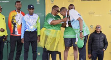 A woman in a yellow skirt awards a medal to a man leaning toward her. Five other people wearing sports attire stand nearby, looking on. The background displays logos and text from a local municipality. The mood is celebratory.