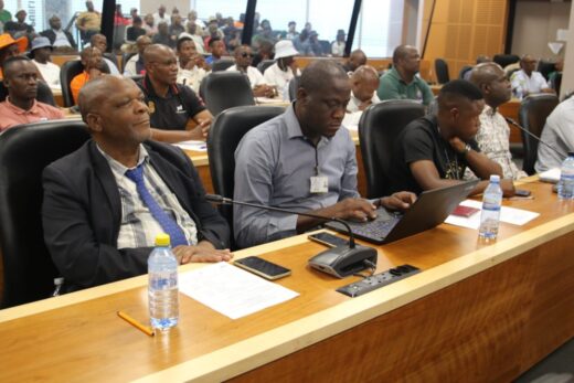 A conference room filled with attendees seated at long tables. Individuals focus on listening and typing on laptops. The atmosphere is serious and attentive.
