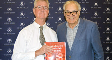Two men are standing smiling in front of a backdrop with "AKADEMIA" logos. One holds a book titled "Konstruktiewe Joernalistiek."