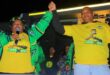 Two people wearing yellow "Vote ANC" shirts hold hands triumphantly. One speaks into a microphone. The background is dark, conveying a lively, energetic atmosphere.