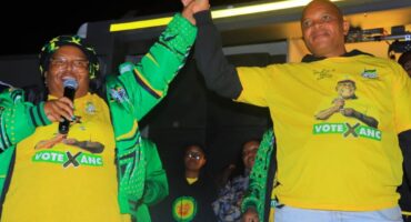 Two people wearing yellow "Vote ANC" shirts hold hands triumphantly. One speaks into a microphone. The background is dark, conveying a lively, energetic atmosphere.