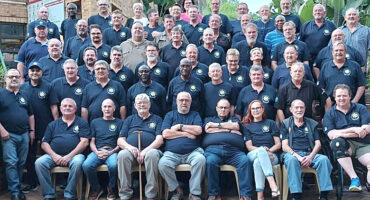 A large group of men and a woman, mostly middle-aged, wearing matching navy shirts with an emblem, pose together outdoors. The mood is friendly and unified.
