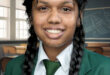 Smiling student in green school uniform with braided hair stands in a classroom. The background has desks, chairs, and a chalkboard with writing.