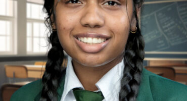 Smiling student in green school uniform with braided hair stands in a classroom. The background has desks, chairs, and a chalkboard with writing.