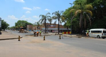 Street construction scene with workers in orange vests and a backhoe under a sunny sky. Palm trees line the road, and a van is parked nearby.