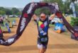 A person triumphantly crosses a triathlon finish line, holding a black banner overhead. Spectators and colorful sponsor signs line the dirt path.