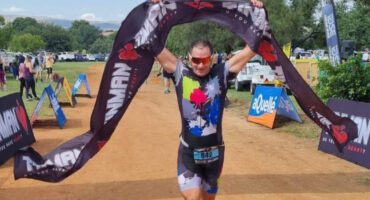 A person triumphantly crosses a triathlon finish line, holding a black banner overhead. Spectators and colorful sponsor signs line the dirt path.