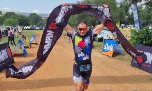 A person triumphantly crosses a triathlon finish line, holding a black banner overhead. Spectators and colorful sponsor signs line the dirt path.