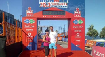 A smiling couple stands under the Ironman 70.3 South Africa finish line arch with a blue sky backdrop. The scene conveys accomplishment and joy.
