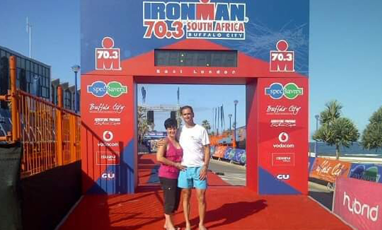 A smiling couple stands under the Ironman 70.3 South Africa finish line arch with a blue sky backdrop. The scene conveys accomplishment and joy.