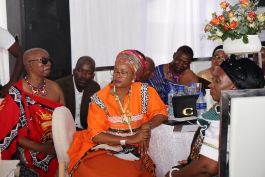A group of people in vibrant traditional attire are seated indoors, engaging in conversation. The mood appears formal and thoughtful, with decorative flowers in the background.