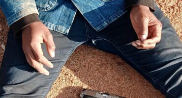 A person in a blue denim jacket and jeans is sitting on sandy ground, legs apart. A handgun rests on the sand between their legs. The scene feels tense.