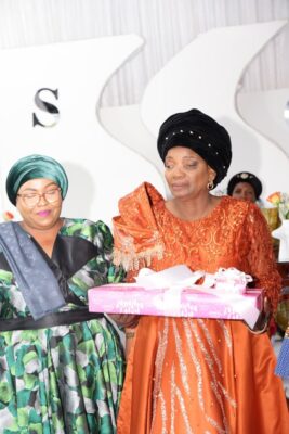 Two women in elegant attire stand together. One wears a green floral dress and teal headwrap, while the other wears an orange dress and black headwrap, holding a pink gift box wrapped with ribbon. The setting appears festive, conveying a celebratory mood.