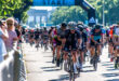 Cyclists in colorful jerseys race from the start line under a clear sky, surrounded by cheering spectators. The mood is energetic and competitive.