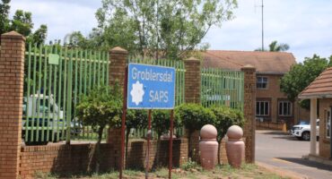 The image shows a sign for Groblersdal SAPS in front of a brick building with a green fence. Trees and potted plants are located in front of the fence.
