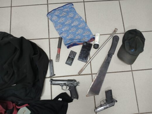 Various items shown on a tiled floor, including two guns, a machete, a screwdriver, mobile phones, a black cap, a shirt, and a package. The scene conveys a sense of tension.