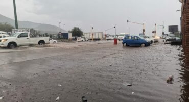 Flooded street with cars navigating through waterlogged intersection under a cloudy sky. Reflections and debris indicate recent heavy rain.