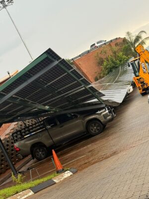 A collapsed solar panel carport leans onto a parked truck, partially damaging the roof. A yellow construction vehicle is beside the scene.