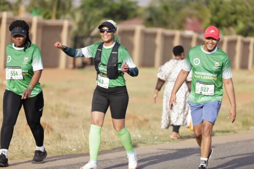 Three smiling runners in green shirts and numbered bibs participate in a marathon. The sunny day enhances their cheerful mood on the rural road.