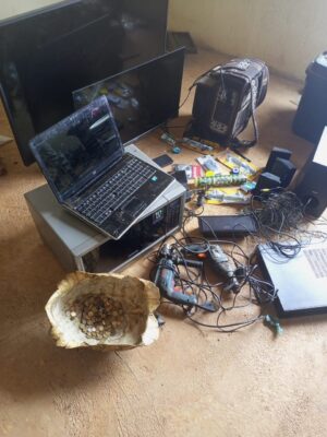 A cluttered room with dusty electronics: two laptops, TV screens, tangled wires, drills, a patterned bag, and a bowl of scattered coins on the floor.