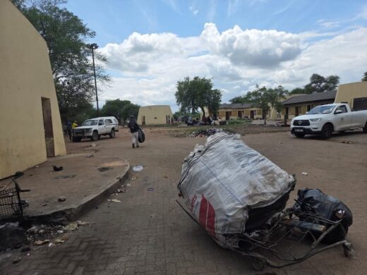 Rural scene with scattered trash and debris. A large white sack on a cart is in the foreground. Cloudy sky above adds a muted, somber tone.