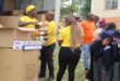 Volunteers in yellow shirts distribute soccer balls to schoolchildren in uniforms. Cardboard boxes are in the background, creating a cheerful atmosphere.