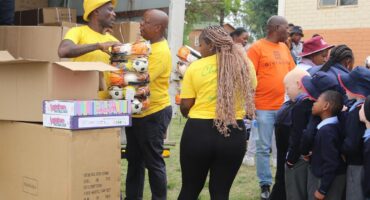 Volunteers in yellow shirts distribute soccer balls to schoolchildren in uniforms. Cardboard boxes are in the background, creating a cheerful atmosphere.