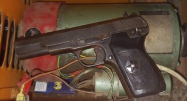 A black pistol with a star emblem on the grip lies on a cluttered yellow shelf. The shelf also contains wires and a cylindrical tool.