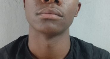 A person wearing a black T-shirt looks directly at the camera with a neutral expression. The background is a plain, light-colored wall.