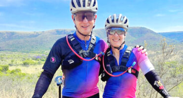 A smiling man and woman wearing matching cycling gear and helmets stand arm in arm. They are outdoors with rolling hills and a clear blue sky behind them.