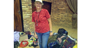 A smiling woman in a red shirt and hat stands indoors near piles of clothes and household items. The setting is a brick-walled room with a relaxed atmosphere.
