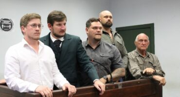 Five men stand in a courtroom, appearing serious and attentive. They are dressed in formal and casual attire, with a wooden railing in front of them.