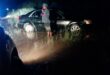 A man stands beside a car stuck in mud at night, illuminated by headlights. Police lights flash in the background, reflecting a tense, uncertain scene.
