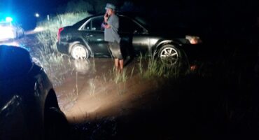 A man stands beside a car stuck in mud at night, illuminated by headlights. Police lights flash in the background, reflecting a tense, uncertain scene.