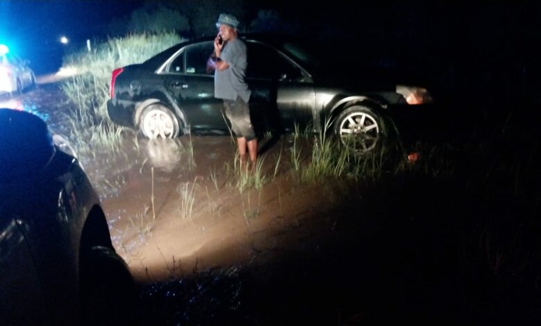 A man stands beside a car stuck in mud at night, illuminated by headlights. Police lights flash in the background, reflecting a tense, uncertain scene.