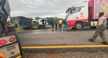 A road scene with a red and white truck and a blue flatbed truck parked on a wet road. Several people in high-visibility gear are conversing closely.