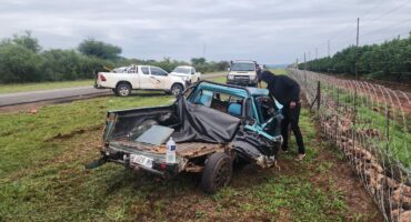 A heavily damaged blue pickup truck lies in a grassy area beside a road, with a person inspecting it. Two white trucks are parked nearby, amidst an overcast sky.