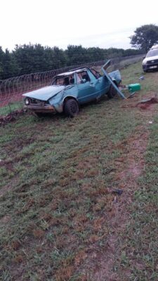 A damaged blue pick-up truck is crashed into a wire fence on grass, with a broken roadside pole beside it. A parked white vehicle is in the background.