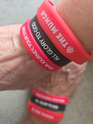 Close-up of wrists wearing three silicone bracelets in red and black. The red reads "THE MUNGA" and "YOUR JOURNEY, YOUR STORY", black reads "ALL GLORY TO GOD".