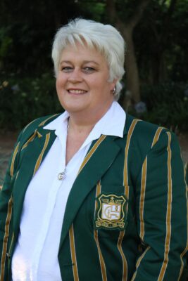 Smiling woman with short white hair wearing a green blazer with yellow stripes and an emblem, standing outdoors in a garden setting with trees.
