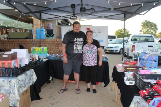 A smiling couple stands under a market stall canopy. The stall displays various colorful items. The atmosphere is lively and welcoming.
