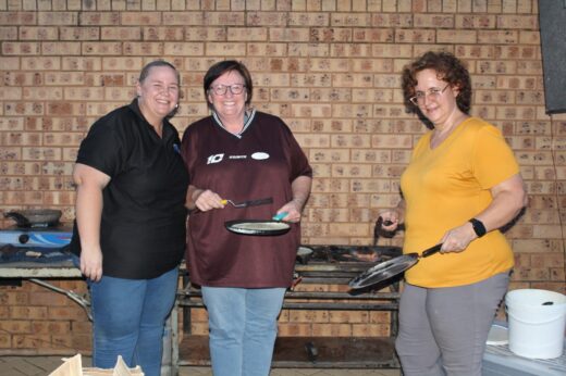 Three people smiling while cooking outdoors. Two hold frying pans, standing in front of a brick wall, creating a friendly and joyful atmosphere.
