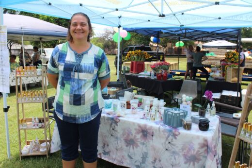 A smiling woman stands at a market stall under a blue canopy. Her display table is adorned with floral products. Sunflowers and balloons are visible in the background, creating a joyful, vibrant atmosphere.